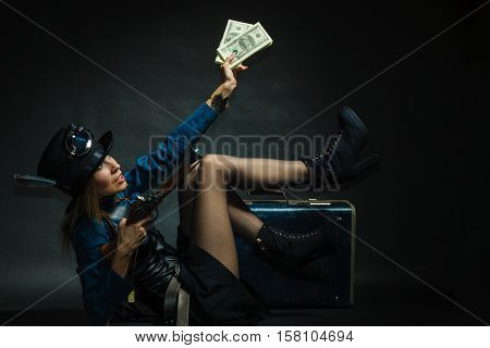 Fashionable subculture weapon concept. Steampunk girl with cash. Young gorgeous lady in victorian fashion lying on floor with banknotes briefcase and blunderbuss.