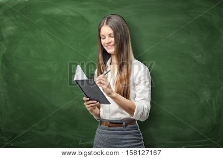 A young businesswoman holding her open day planner and a pen and going to write something on the green chalkboard background. Daily planning. Time-management. Secretary work.