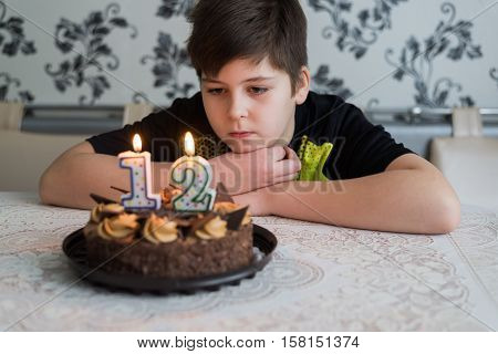 Teen boy looks thoughtfully at a cake with candles on the twelfth day of birth