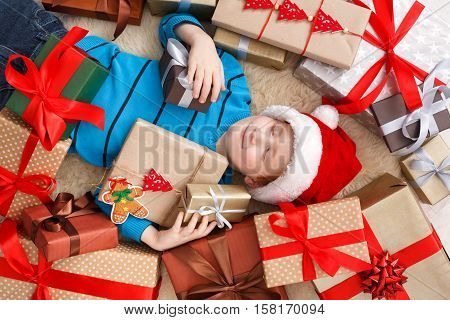 Happy boy child in santa hat lay on warm carpet among Christmas gift boxes. Xmas holiday concept. Top view of excited cute kid got lots of presents