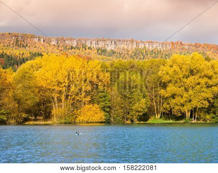 Sandstone rock formation in the middle of colorful autumn forest. Water surface on the foreground. Dramatic evening view. Tisa Rocks, aka Tiske Walls, Czech-Saxon Switzerland, Czech Republic