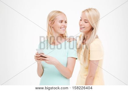 Picture of young attractive lady showing her phone to the friend. Isolated over white background. Looking at each other.