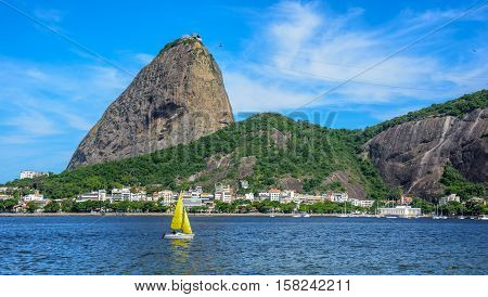 Yellow small sailing yacht and classic daytime scenic profile view of Sugarloaf Mountain, Pao de Acucar, standing above Botafogo Bay in Rio de Janeiro, Brazil