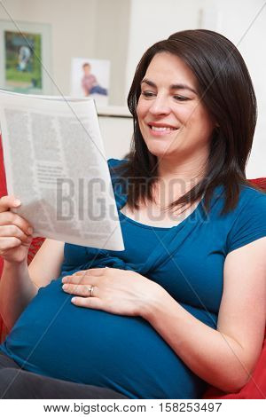 Pregnant Woman Sitting On Sofa Reading Magazine