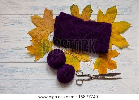 Violet yarn knit fabric wooden knitting needles scissors and yellow fallen leaves are on white vintage wooden desk. Top view.