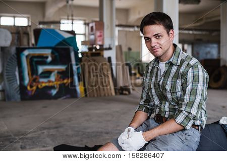Handsome carpenter in protective glasses is looking at camera and smiling while standing near his wooden object in the workshop.