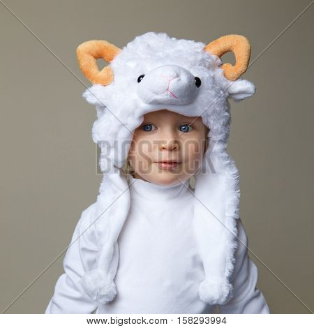 Cute adorable pretty Caucasian baby toddler with large blue eyes wearing a sheep hat hood with yellow horns on top and white shirt standing on a light background New Year concept studio