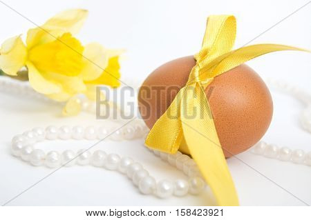 Easter egg with yellow ribbon lies on white background beside pearl necklace and narcissus.