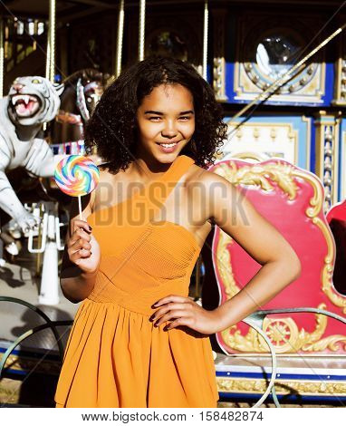 cool real modern teenage girl with candy near carousels at amusement park walking, having fun