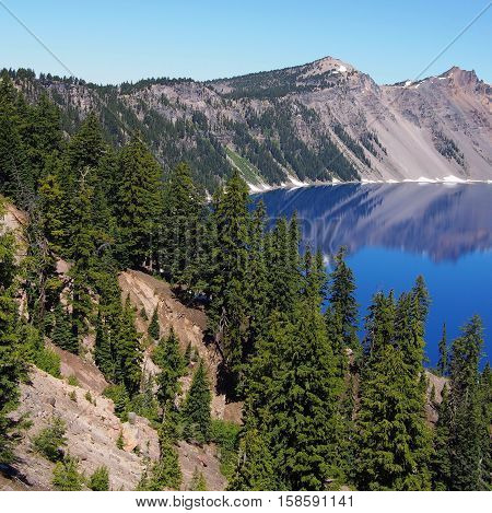 The rim of Crater Lake reflects in the deep blue waters of the lake on a sunny summer day.