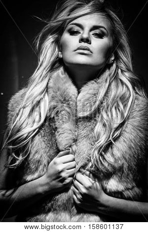 Fashion portrait of young blond woman in fur coat posing on dark background