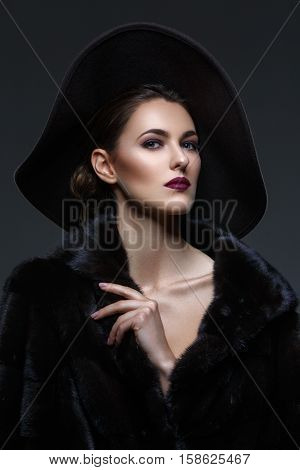 Beautiful young woman with glamorous make-up in luxurious fur coat and fashion hat on black background. Copy space.