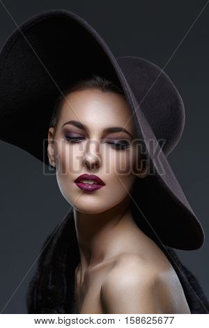 Beautiful young woman with glamorous make-up in luxurious fur coat on naked body and fashion hat on black background. Copy space.