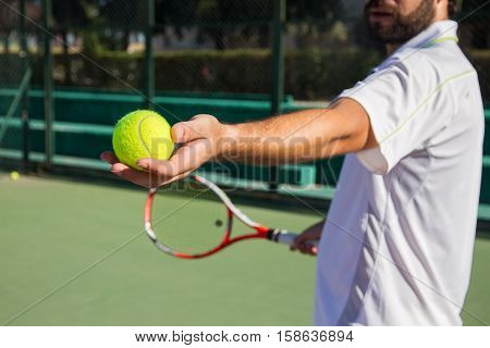 Professional tennis player, bearded young man offering tennis ball to play the game. He has a racket in her right hand and wears a white cap.