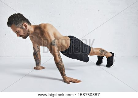 Strong brutal tattoed and ripped male athlete shows how to do pseudo push-ups, isolated on white spacious room, top position