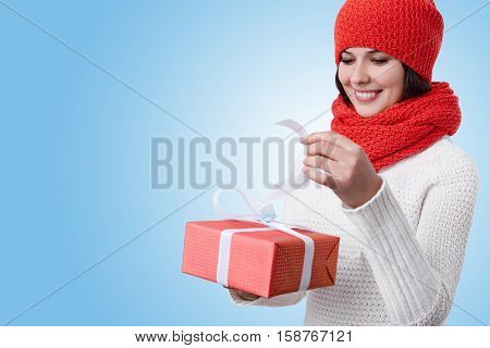 New Year Time. A joyful woman wearing white sweater and red hat with scarf having a pleasant smile and being happy from receiving and unpacking presents. A happy pretty woman having winter holidays