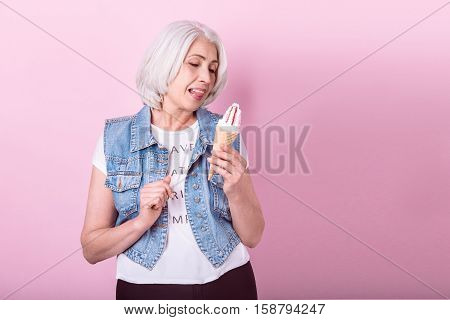 So tasty. Beautiful senior woman licking her lips and holding an icecream while standing against isolated pink background.