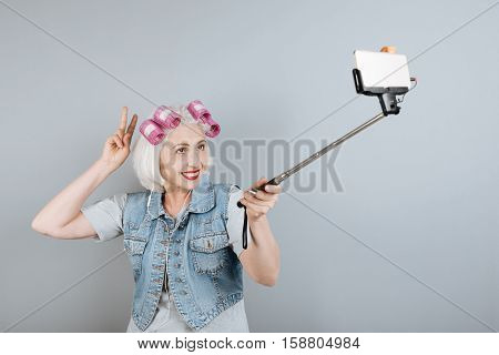 Modern technologies. Cheerful senior beautiful woman using headphones and taking selfie while standing against isolated gray background.