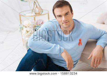 The red ribbon. Handsome positive delighted man leaning on the sofa and wearing the red ribbon while taking part in the international AIDS day