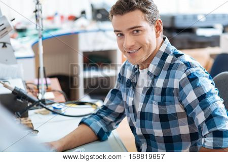 Repair process. Young handsome ambitious man sitting in a workroom while smiling and repairing drones