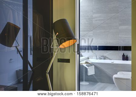 Orange glowing design lamp on the background of the entrance to the bathroom in a modern style. Bathroom tiled with gray and white tiles. There is a bath, toilet and bide, stand with towel and book.