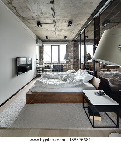 Bedroom in a loft style with brick wall and concrete ceiling. There is a TV, bed with pillows, lamps with lampshades, wardrobe with glass sliding doors, tables, armchair, carpet on the floor.