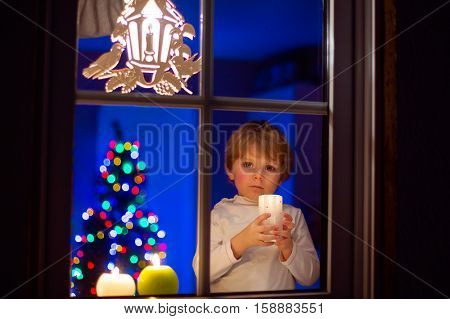 Beautiful little kid boy, standing by window at Christmas time and holding candle. Child waitng for Santa. With colorful lights from Christmas tree on background.