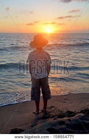 Small boy stands on the beach at the sundown, hands in the trouser pockets and looks as the sun