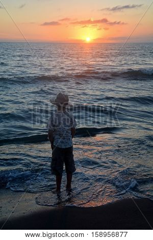 Small boy stands on the beach at the sundown, hands in the trouser pockets and looks to the horizon