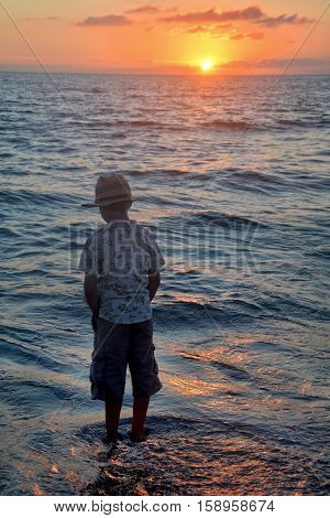 Small boy stands on the beach at the sundown, hands in the trouser pockets and looks to the horizon