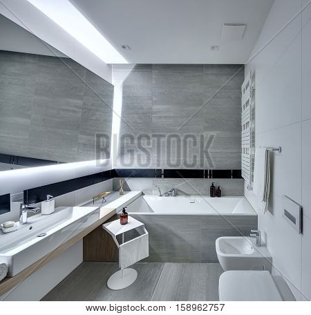 Modern bathroom tiled with the white and gray tiles. There is a white sink with a soap, large mirror, bath, towel holder, towel radiator, toilet and a bide, stand with a towel and a pump bottle.