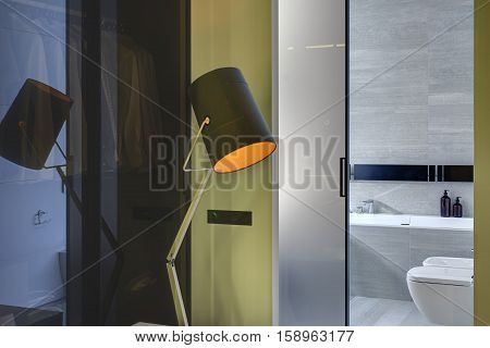Design glowing lamp on the background of the entrance to the modern bathroom with a glassy door. Bathroom tiled with gray and white tiles. There is a white bath, white toilet and a bide. Horizontal.