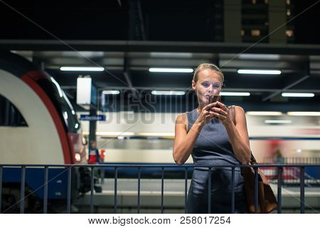 Pretty, young female commuter waiting for her daily train in a modern trainstation, using her cellphone while waiting (color toned image)