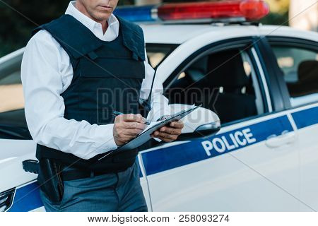 Partial View Of Middle Aged Male Police Officer In Bulletproof Vest Writing In Clipboard Near Car At
