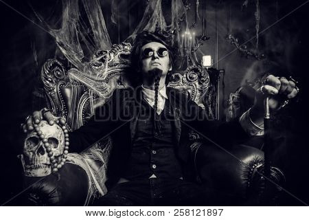 Halloween. Frightening gloomy man in a black tailcoat sits in an armchair with a skull in an old abandoned castle. The Dark Lord. Vampire man. 