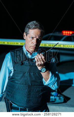 Serious Mature Policeman In Bulletproof Vest Talking On Radio Set Near Car At Crime Scene