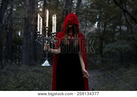 Mystic Girl In Red Cloak And Hood Holding Candelabrum With Candles In Dark Forest
