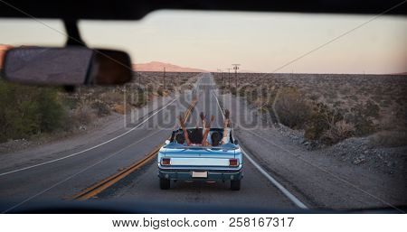 Group Of Friends On Road Trip Driving Classic Convertible Car Viewed Through Windshield Of Following Vehicle