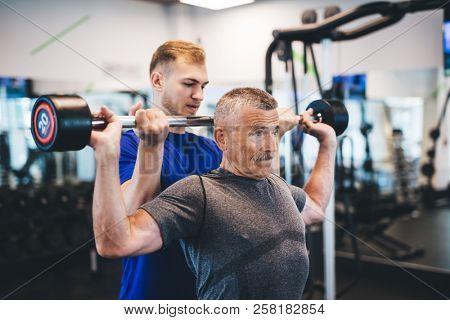 Senior man lifting weights with help of gym assistant. Work of personal trainer. Bodybuilding.
