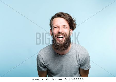 Happiness Enjoyment And Laugh. Man With A Wide Grin. Portrait Of A Young Bearded Hipster Guy On Blue