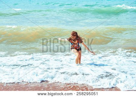 A Young Girl In Shorts Stands With Her Back Against The Background Of Big Waves