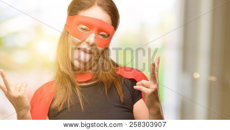 Middle age super hero woman wearing red mask and cape looking at camera showing tong and making victory sign with fingers