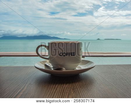Cup Of Coffee On A Saucer Placed On A Wooden Table With A Tropical Seascape In The Background.  The 