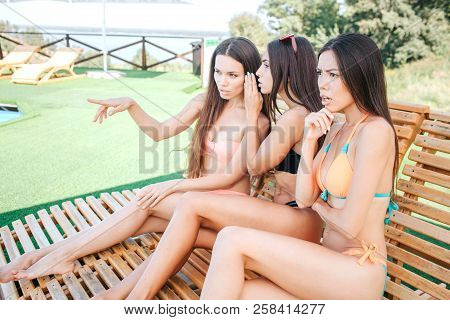 Amazed And Serious Beautiful Young Women Sit On Sunbeds. First One Is Pointing Forward. Second One I