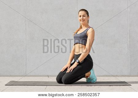 Satisfied Smiling Woman Being In Good Mod After Doing Crunches On Fitness Mat, Stands On Knees, Dres
