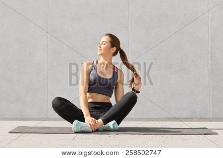 Relaxed Beautiful Caucasian Woman Holds Pony Tail, Sits Crossed Legs On Fitness Mat, Being In High S