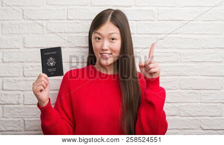 Young Chinese woman over brick wall holding passport of America surprised with an idea or question pointing finger with happy face, number one
