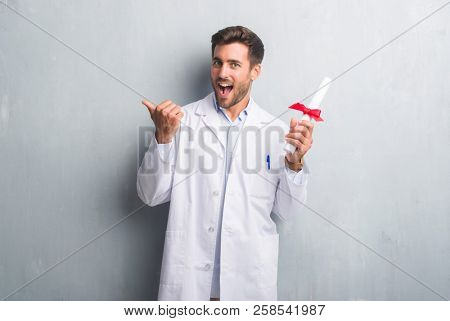 Handsome young doctor man over grey grunge wall holding diploma pointing and showing with thumb up to the side with happy face smiling