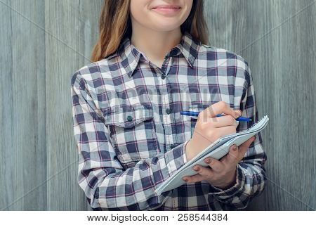 A Portrait Of A Young Smiling Student In Checkered Shirt Writing Some Notes In Her Notepad
