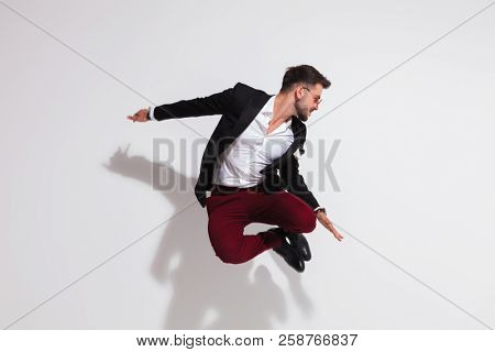 smart casual man jumping and doing a heel click near a white wall while looking down to side, full body picture